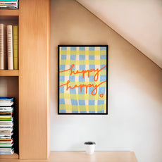 Framed hand-painted gingham artwork with 'happy happy' text on a wall next to a bookshelf