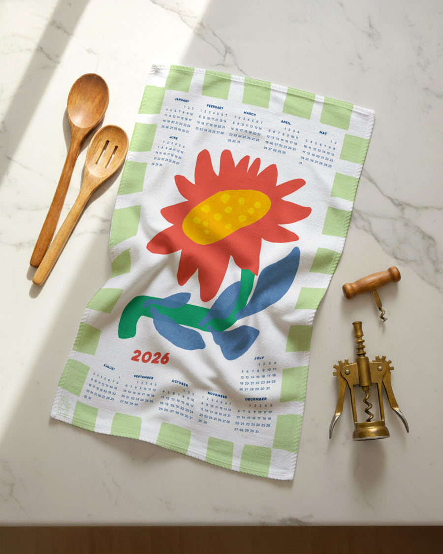 Flat lay of 2026 Sunflower Calendar Tea Towel with ochre center and green striped border, shown with kitchen utensils on a marble surface