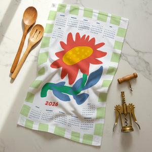 Flat lay of 2026 Sunflower Calendar Tea Towel with ochre center and green striped border, shown with kitchen utensils on a marble surface