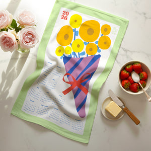 Flat lay of 2026 Vintage Bouquet Calendar Tea Towel featuring yellow and blue floral bouquet with seafoam border, on a marble countertop with fresh flowers and beautiful food.