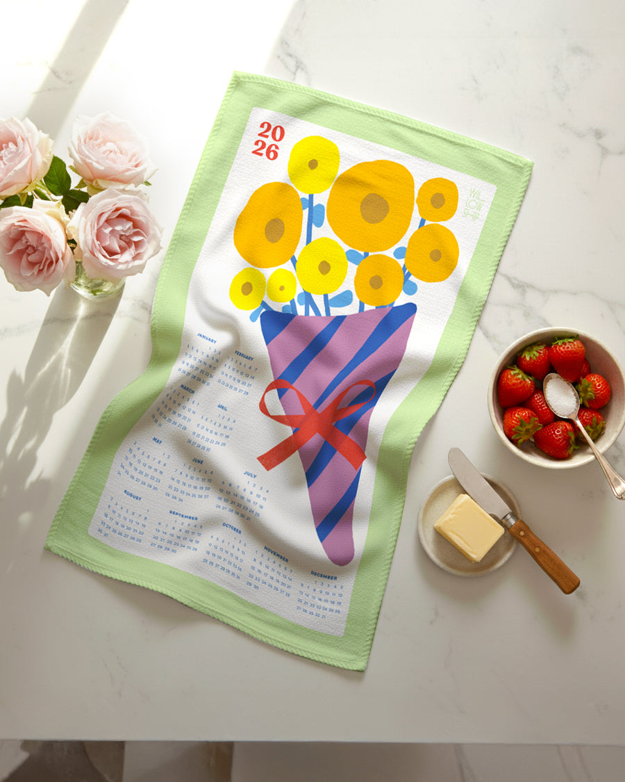 Flat lay of 2026 Vintage Bouquet Calendar Tea Towel featuring yellow and blue floral bouquet with seafoam border, on a marble countertop with fresh flowers and beautiful food.