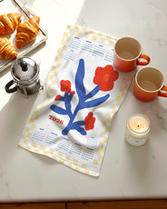 Flat lay of 2026 Red Flowers Calendar Tea Towel featuring red blooms with butter-yellow check border, shown on a marble countertop with croissants, coffee, and a candle.