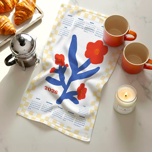 Flat lay of 2026 Red Flowers Calendar Tea Towel featuring red blooms with butter-yellow check border, shown on a marble countertop with croissants, coffee, and a candle.