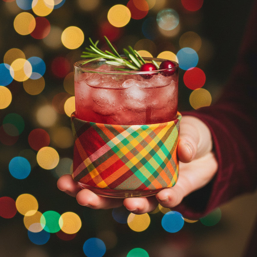 Close-up of a hand holding a holiday cocktail wrapped in a Merry Madras cocktail napkin, with colorful Christmas bokeh lights in the background.