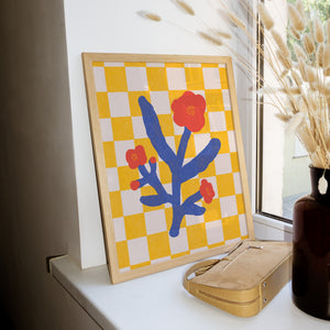 Framed abstract art with blue and red design on a yellow checkered background, placed on a surface next to a suitcase and vase.