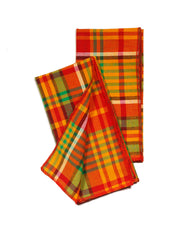 Two Merry Madras dinner napkins partially folded on a white background, showing the bold red, green, and warm yellow plaid pattern with red serged edges.