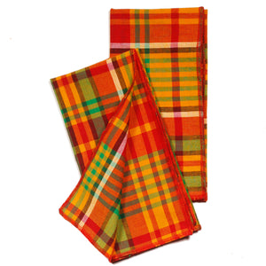 Two Merry Madras dinner napkins partially folded on a white background, showing the bold red, green, and warm yellow plaid pattern with red serged edges.